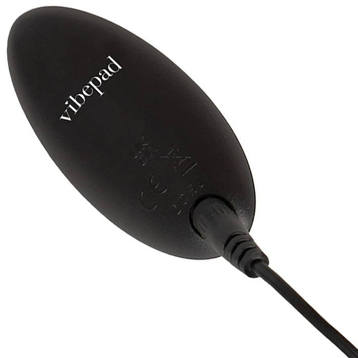 A black, oval-shaped device labeled "VibePad 3 Ride On Hands-Free Humping Vibrator with G-Spot Probe" by Orion, featuring a connected cable on one end. The surface showcases small printed symbols and text indicating certification and specifications, suggesting it is a vibrating pad electronic accessory.