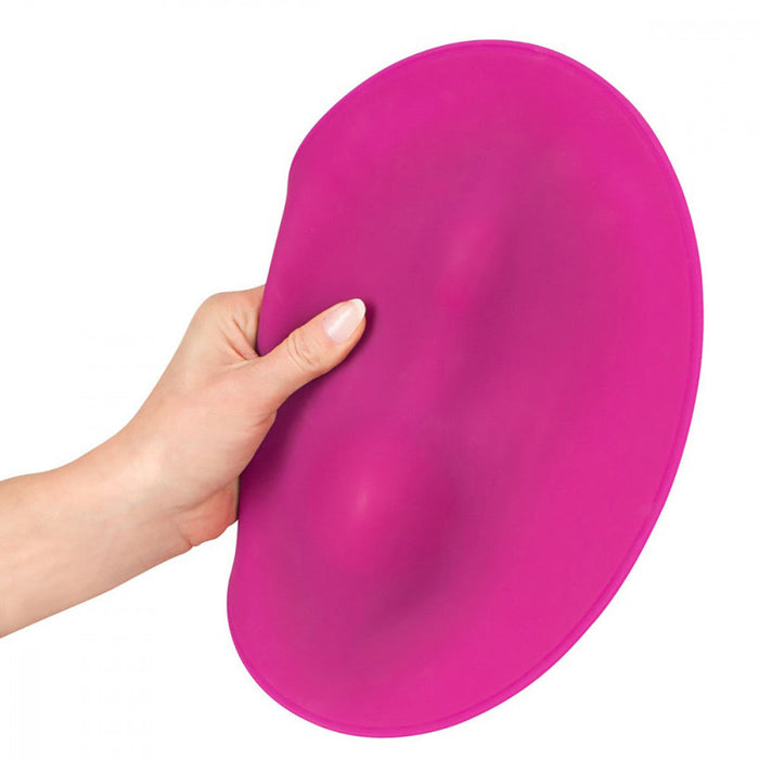 A hand is holding a flexible, flat, round magenta object against a white background. The object appears to be the VibePad Ride On Hands-Free Humping Vibrator made of a rubber-like material from the brand Orion.