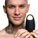 A smiling person with a shaved head and tattoos on their neck holds the XR Brands Vibrating Silicone Waterproof Ball Sack, a black handheld device with a loop handle. The device appears to be vibrating, indicated by blue wavy lines on either side.