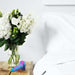 A clear glass vase containing white hydrangeas and green berries is placed on a wooden bedside table next to a bed with white linens. The table also holds the colorful, striped Vibrotize 10 Function Vibrating Silicone Butt Plug by Blush.