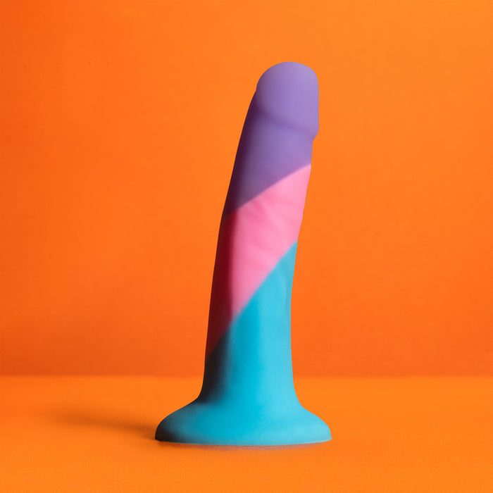 The Vision of Love Silicone Suction Cup 5.5 Inch Dildo by Blush stands upright against an orange background, showcasing its vibrant and colorful design. Crafted from platinum silicone, this beginner dildo features shades of blue, pink, and purple arranged in diagonal sections. It also comes equipped with a suction cup base for hands-free fun.