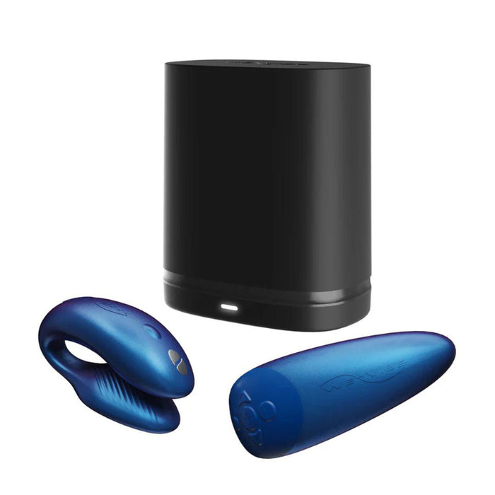 A black rectangular device with a small indicator light near the bottom sits in front of two blue accessories: the We-Vibe Chorus Remote & App Controlled Couples' Vibrator, featuring a contoured C-shape, and another ergonomically designed cylindrical item with textured surface and touch-sensitive vibrations. Both items are adorned with subtle embossed detailing.