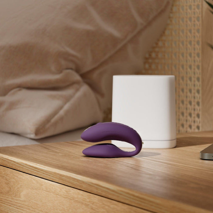 A We-Vibe Chorus Remote & App Controlled Couples' Vibrator in vibrant purple, featuring an adjustable fit and touch-sensitive vibrations, is placed on a wooden nightstand. In the background, a neatly made bed with white pillows and a beige headboard can be seen. The nightstand also holds a white square lamp and a sleek, metallic object.