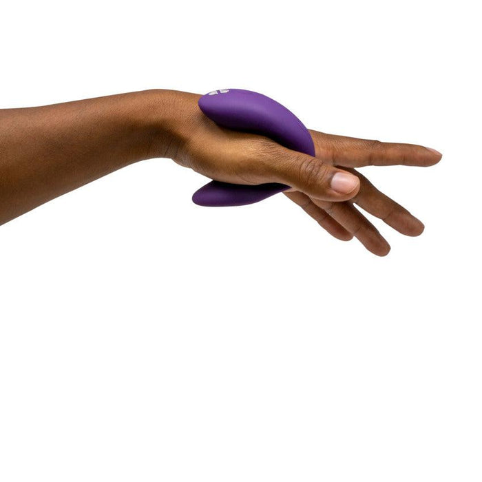 A close-up of a hand holding the We-Vibe Chorus Remote & App Controlled Couples' Vibrator in purple, which features a smooth material and an adjustable fit design with buttons. The background is white, emphasizing the hand and the object.