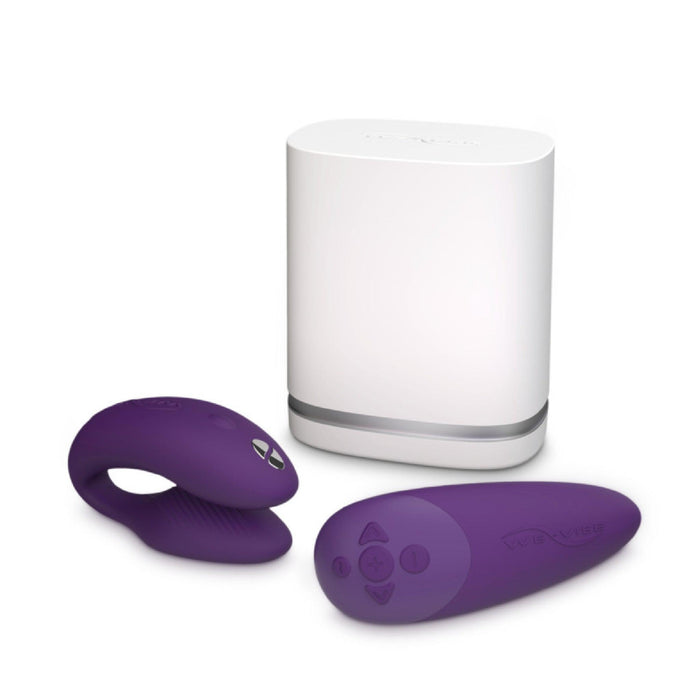 A white rectangular charger stands behind two purple silicone devices from the We-Vibe brand: the We-Vibe Chorus Remote & App Controlled Couples' Vibrator, which offers an adjustable fit and touch-sensitive vibrations. To the left is a ring-like device, while on the right is a remote control with three buttons. These items are designed for intimate use.