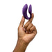 In a plain white background setting, a hand holds the purple We-Vibe Chorus Remote & App Controlled Couples' Vibrator by We-Vibe. The fingers are positioned to display the device, showcasing its curved and ergonomic design for an adjustable fit and touch-sensitive vibrations.