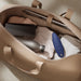 An open beige leather bag on a beige carpet holds a black water bottle, the We-Vibe Melt Rechargeable Pleasure Air Clitoral Stimulator in blue, a smartphone with the We-Connect™ app, a folded shirt, and a laptop. The bag features two handles and is lined with brown fabric.