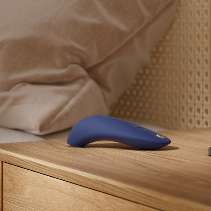 A sleek, blue We-Vibe Melt Rechargeable Pleasure Air Clitoral Stimulator rests on a wooden nightstand next to a bed with a woven headboard and beige pillows. The device features buttons on the top surface and boasts contoured lines designed for clitoral air pulse stimulation. The scene is softly lit, suggesting a cozy bedroom setting.