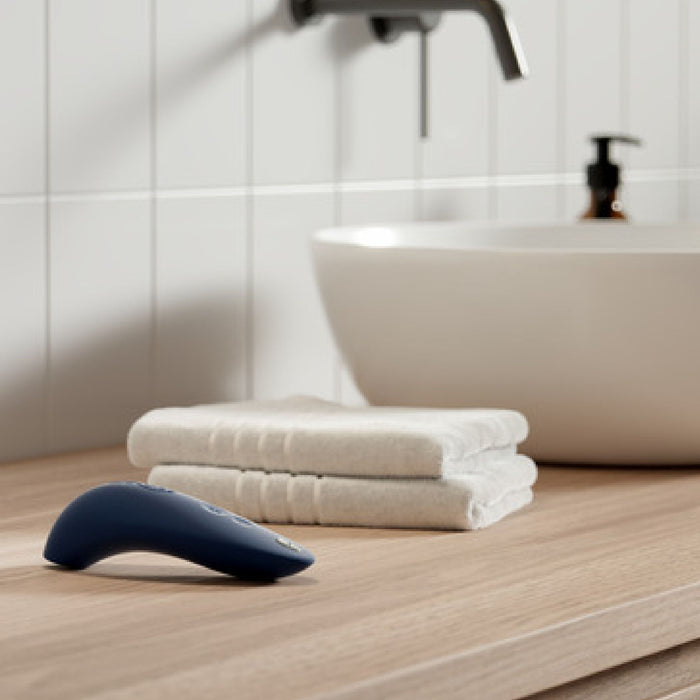 A modern bathroom sink with a sleek silver faucet is shown. On the wooden countertop in front of the sink, there are two neatly folded white towels and a We-Vibe Melt Rechargeable Pleasure Air Clitoral Stimulator in dark blue by We-Vibe. The background features white tiled walls and a black pump soap dispenser.