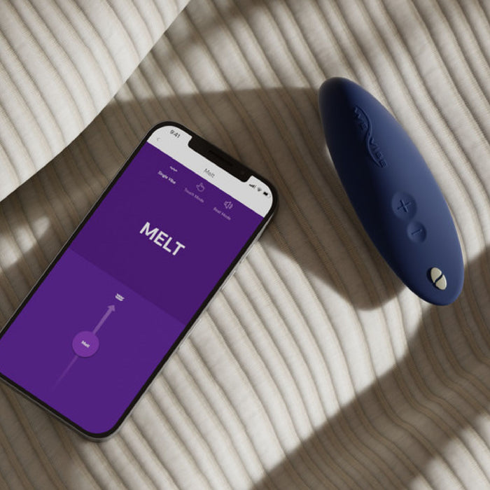 A smartphone displaying a purple app screen with the word "MELT" is placed on a striped surface beside the We-Vibe Melt Rechargeable Pleasure Air Clitoral Stimulator in blue. Both items are well-lit with natural light casting soft shadows.