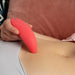 A close-up of a person holding the We-Vibe Melt Rechargeable Pleasure Air Clitoral Stimulator - Coral against their abdomen. The device, designed for clitoral air pulse stimulation, is curved and appears to be made of a soft, rubbery material. The person is lying down and partially visible, wearing light-colored clothing.