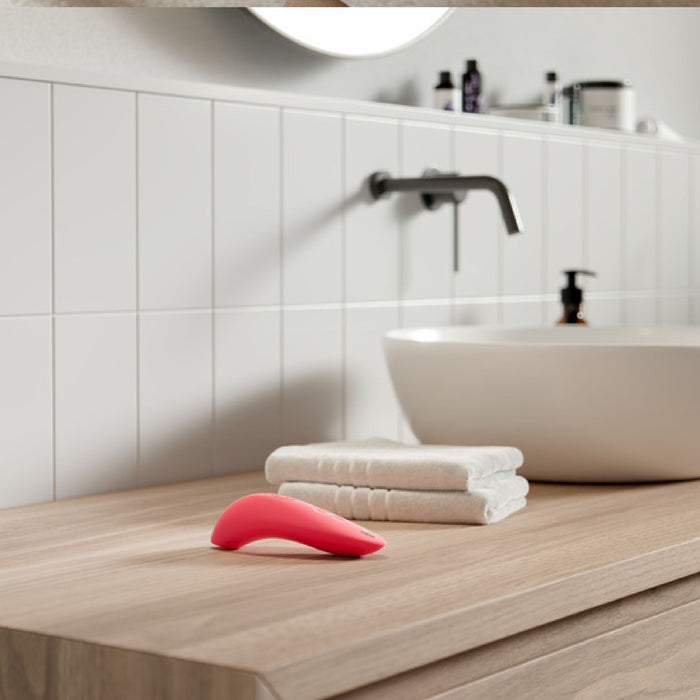 A modern bathroom counter features a white sink, a sleek black faucet, and an array of toiletries in the background. In the foreground, two folded white towels are placed on the wooden countertop alongside a We-Vibe Melt Rechargeable Pleasure Air Clitoral Stimulator in Coral. The wall behind is adorned with pristine white tiles.