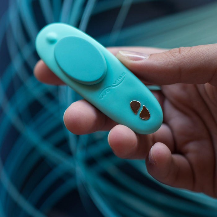 A person holds a small, teal electronic device with an oval button and two metallic contacts near the tip, resembling the sleek We-Vibe Moxie + Hands-Free Remote or App Controlled Panty Vibrator. The background is blurred with swirls of blue and green lines.