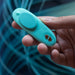 A person holds a small, teal electronic device with an oval button and two metallic contacts near the tip, resembling the sleek We-Vibe Moxie + Hands-Free Remote or App Controlled Panty Vibrator. The background is blurred with swirls of blue and green lines.