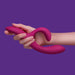 A person's hand holding the We-Vibe Nova 2 Powerful Flexible G-Spot Rabbit Vibrator, featuring its pink, curved silicone design with two prominent ends—one larger and bulbous and the other slimmer and curved—against a solid purple background.