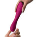 Two hands holding a fuchsia-colored, curved We-Vibe Rave 2 Powerful Adjustable G-Spot Vibrator with button controls visible on the side. One hand, with a light pink manicure, gently receives the app-enabled massager from the other hand. The background is plain white.