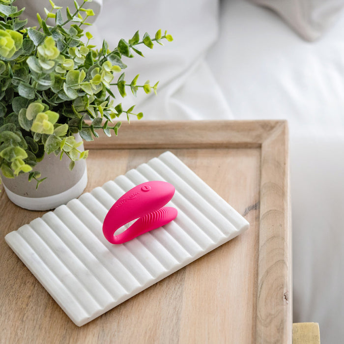 The We-Vibe Sync Lite App Controlled Wearable Couples Vibrator in pink, featuring textured silicone grips, is positioned on a ribbed white ceramic tray on a wooden surface. Next to it stands a potted plant with green leaves and behind them, a section of a neatly made white bed is visible.