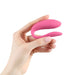 A hand with light skin is holding a pink, U-shaped We-Vibe Sync Lite App Controlled Wearable Couples Vibrator against a white backdrop. The smooth and flexible device, by the renowned brand We-Vibe, is designed for intimate use. Adding an elegant touch to the scene, the person’s fingernails are manicured with light pink polish.