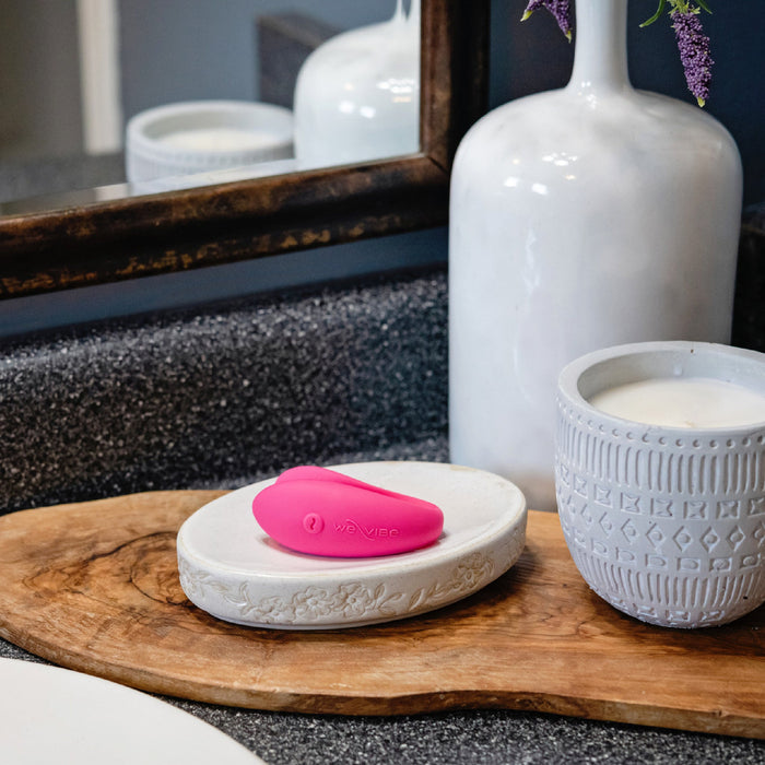 A modern bathroom counter with a granite surface showcases a white vase with flowers, a patterned white jar, and a wooden tray holding the We-Vibe Sync Lite App Controlled Wearable Couples Vibrator in pink. A mirror is partially visible in the background.