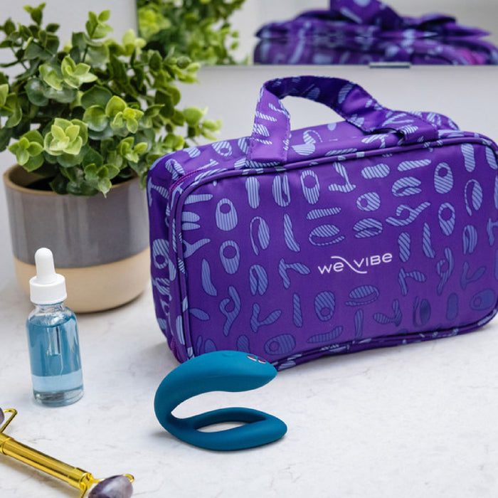 A purple patterned We-Vibe cosmetic bag is placed on a bathroom counter. Beside it are a blue dropper bottle with liquid, a facial roller, and a potted plant. A green We-Vibe Sync O Hands-Free Wearable Couples Vibrator lies in front of the bag. A mirror reflects part of the scene.