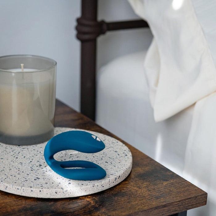 A small, curved green We-Vibe Sync O Hands-Free Wearable Couples Vibrator by We-Vibe rests on a round, speckled stone coaster next to an unlit gray candle on a wooden bedside table. A neatly made bed with white sheets is partially visible in the background, hinting at a serene space perfect for using app-controlled wearable couples vibes.