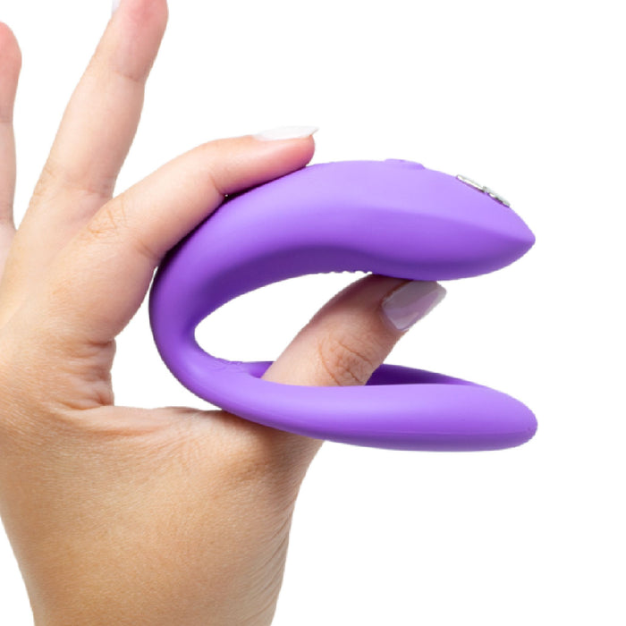 A hand holds the We-Vibe Sync O Hands-Free Wearable Couples Vibrator - Purple with two fingers through its open ends. The device from We-Vibe showcases a smooth, sleek design and appears to be made of silicone. This waterproof accessory is perfect for shared moments. The background is white.