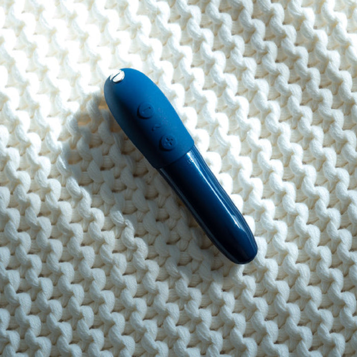 The We-Vibe Tango X Powerful Bullet Vibrator in blue, a sleek cylindrical device with buttons, rests on a textured, white knitted surface. The waterproof vibrator is illuminated from the top left, casting a soft shadow below and to the right.