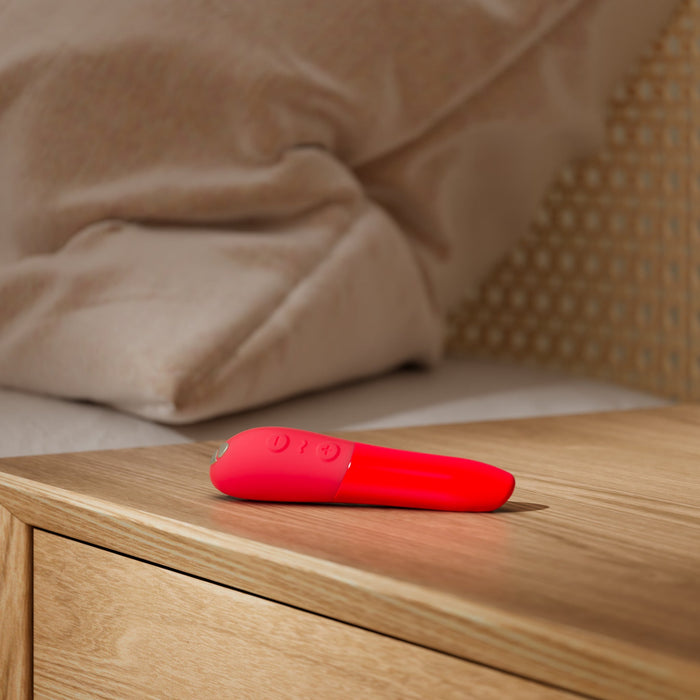 The We-Vibe Tango X Powerful Bullet Vibrator in a vibrant red color, probably used as a personal massager, sits quietly on a wooden bedside table. This whisper-quiet device is USB rechargeable. In the background, you can see a beige pillow and part of a bed with a woven headboard.