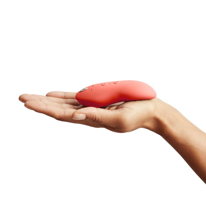 A person holds a We-Vibe Touch X Lay On Vibrator - Coral, a small, curved, pink device made of premium silicone, in their hand against a white background. The device features buttons on its surface, indicating it is an electronic gadget or personal care item from the brand We-Vibe.