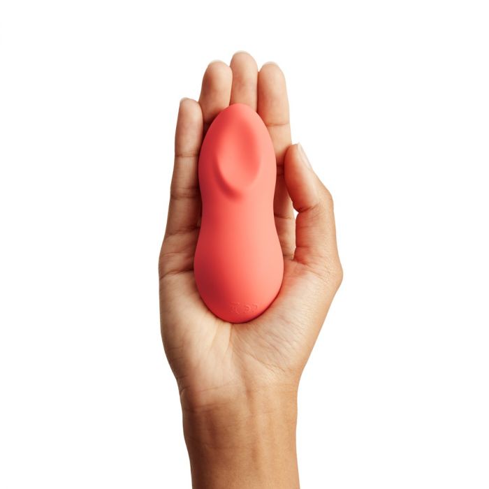 A hand holding a small, smooth, coral-colored We-Vibe Touch X Lay On Vibrator with a curved indentation. Made of premium silicone and USB rechargeable, this ergonomic device fits comfortably in the palm. The white background highlights its shape and color.