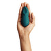 A hand holding a small, green-colored, ergonomically designed, waterproof We-Vibe Touch X Lay On Vibrator with a silicone coating against a white background. The vibrator is oval-shaped with a slightly curved indentation on top.