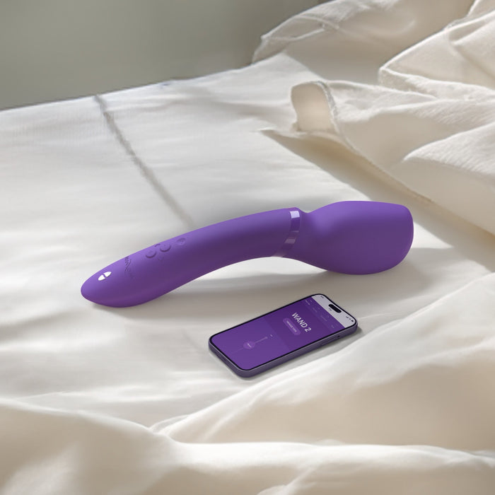 On a white bed, near a smartphone displaying an app with purple visuals and a temperature symbol, lies the We-Vibe Wand 2, a powerful cordless waterproof vibrator by We-Vibe in purple, equipped with buttons and resembling a wand.