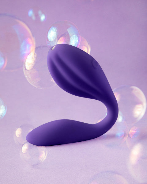 A sleek, purple, ergonomically designed object set against a soft purple background with iridescent bubbles floating around. The Blush Wellness Duo Hands Free Wearable and Couples Vibrator features a curved shape and vibration functions with smooth, flowing lines, giving it a modern and sophisticated appearance.