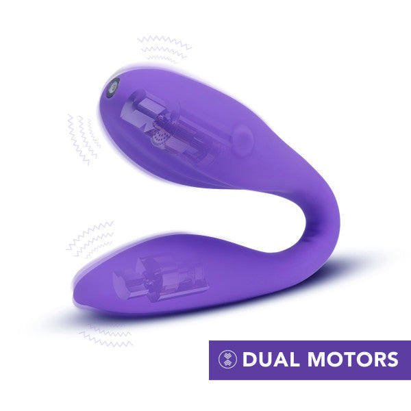 A Wellness Duo Hands Free Wearable and Couples Vibrator by Blush, showcasing a flexible design in purple with dual motors and Puria™ silicone. The device is depicted vibrating, indicated by wavy lines around it. A small control button is visible on one end, with a caption "DUAL MOTORS" in a purple box at the bottom right corner.