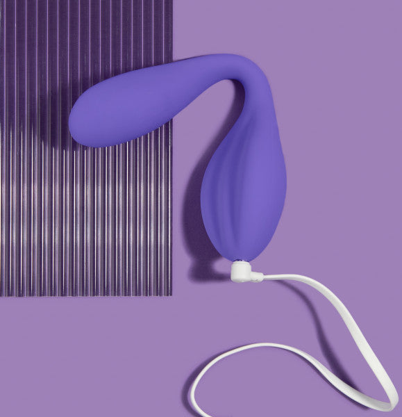 A **Wellness Duo Hands Free Wearable and Couples Vibrator** by **Blush**, featuring a purple, curved, balloon-like silicone design with a long white cord, rests on a flat purple surface beside a vertically striped purple sheet. The background and items exhibit varying shades of purple, forming a cohesive monochromatic aesthetic.