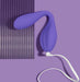 A **Wellness Duo Hands Free Wearable and Couples Vibrator** by **Blush**, featuring a purple, curved, balloon-like silicone design with a long white cord, rests on a flat purple surface beside a vertically striped purple sheet. The background and items exhibit varying shades of purple, forming a cohesive monochromatic aesthetic.