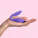 A hand holding the Blush Wellness Duo Hands Free Wearable and Couples Vibrator in purple against a light pink background. The sleek, curved, dual-ended silicone device is designed with one end resembling a bulb and the other featuring a flat, elongated shape.