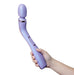 A hand is holding the Wellness Eternal Slim Ergonomic Powerful Remote Control Wand by Blush in a beautiful lavender color. The wand-style personal massager features an elongated handle and a rounded head. It is waterproof and has a couple of visible buttons on the side, likely for controlling its powerful vibrations.