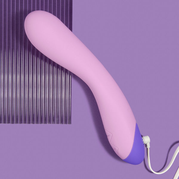 A Wellness G Curve Rumbly Waterproof Silicone G-Spot Vibrator by Blush, featuring a pink and purple design with a wired remote, lies on a purple background. Crafted from silky silicone, the top portion of the background showcases vertical grooves that contrast with the smooth texture beneath the device.