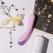 On a white surface, the Wellness G Curve Rumbly Waterproof Silicone G-Spot Vibrator by Blush in pastel pink and purple rests beside a tall white candle adorned with star patterns. Additional decorative items such as crystals and an ornamental sequin string lend a whimsical ambiance. A photograph featuring candlelit lanterns is partially visible nearby.