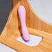 A pastel pink and purple Wellness G Curve Rumbly Waterproof Silicone G-Spot Vibrator by Blush stands upright in a corner of a bamboo shelf with a hexagonal tile background.