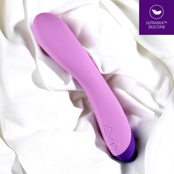 A smooth, purple Wellness G Curve Rumbly Waterproof Silicone G-Spot Vibrator from Blush rests on a white fabric background. With its sleek and curved design perfect for G-spot exploration, the device features control buttons near the base. A purple icon in the top right corner reads "ULTRASILK™ SILICONE".