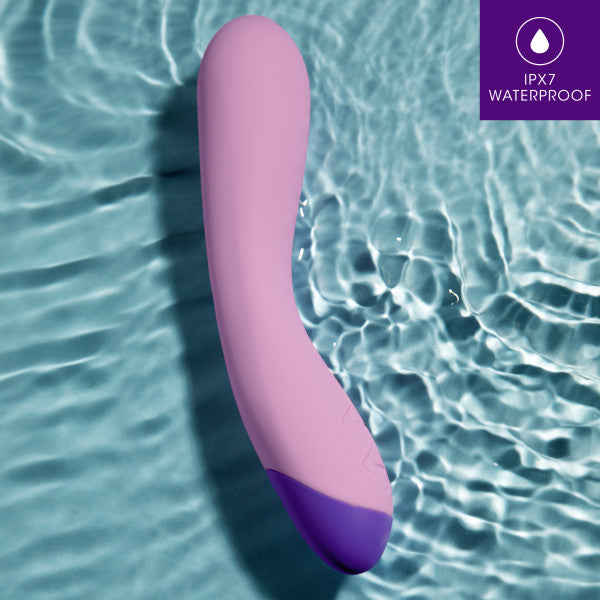 A purple and lavender Wellness G Curve Rumbly Waterproof Silicone G-Spot Vibrator from Blush, made of silky silicone, is submerged in water to showcase its waterproof feature. The upper right corner displays an icon and text stating "IPX7 WATERPROOF" on a purple background, ideal for G-spot exploration.