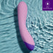 A purple and lavender Wellness G Curve Rumbly Waterproof Silicone G-Spot Vibrator from Blush, made of silky silicone, is submerged in water to showcase its waterproof feature. The upper right corner displays an icon and text stating "IPX7 WATERPROOF" on a purple background, ideal for G-spot exploration.