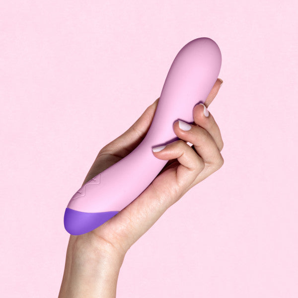 A hand holding the Blush Wellness G Curve Rumbly Waterproof Silicone G-Spot Vibrator against a light pink background. The silky silicone massager has a smooth surface with a wider, rounded end and a set of controls on the lower part.