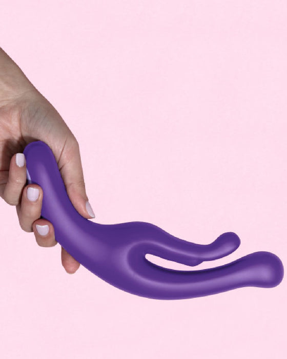 A hand holding a purple, curved, dual-ended silicone **Wellness G Wave Waterproof Rabbit Vibrator with Ergonomic Handle** by **Blush** with powerful 7 function motors against a light pink background.