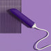 The Blush Wellness Power Bullet - Rumbly Waterproof Ultra Strong Vibrator, connected to a white charging cable, is set against a purple background. The sex toy is partially resting on a textured, translucent surface with vertical ridges.