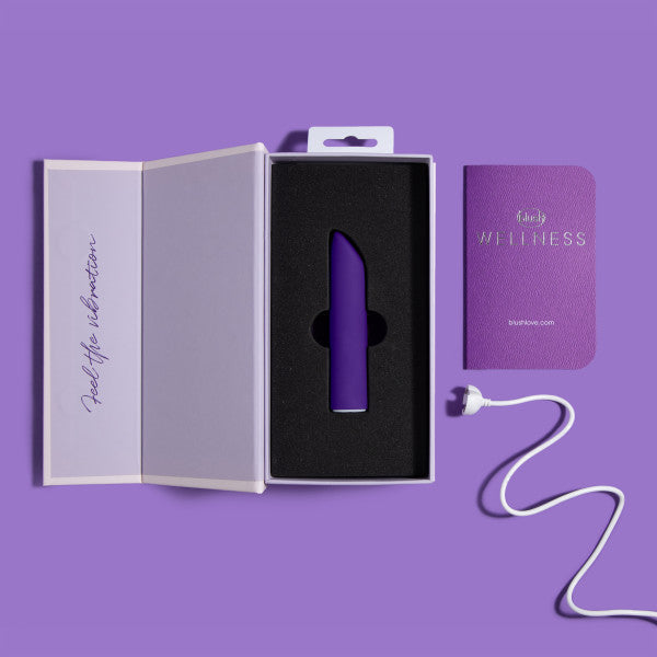 The Wellness Power Bullet - Rumbly Waterproof Ultra Strong Vibrator by Blush, in purple, rests in black foam inside an open white box. To the right, a small purple booklet labeled "Wellness" and a white charging cable are visible. The background is a matching shade of purple. This sleek and waterproof sex toy promises a powerful vibration experience for personal wellness.