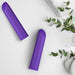 Two "Wellness Power Bullet - Rumbly Waterproof Ultra Strong Vibrators" by Blush, with their sleek purple cylindrical design and rounded ends, are placed on a white surface near neatly folded white towels and sprigs of greenery.