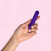 A hand with light skin holds the Blush Wellness Power Bullet - a small, cylindrical, purple vibrator with a rounded end, against a light pink background. The USB rechargeable device is showcased by the person whose neatly manicured fingernails are painted light pink.